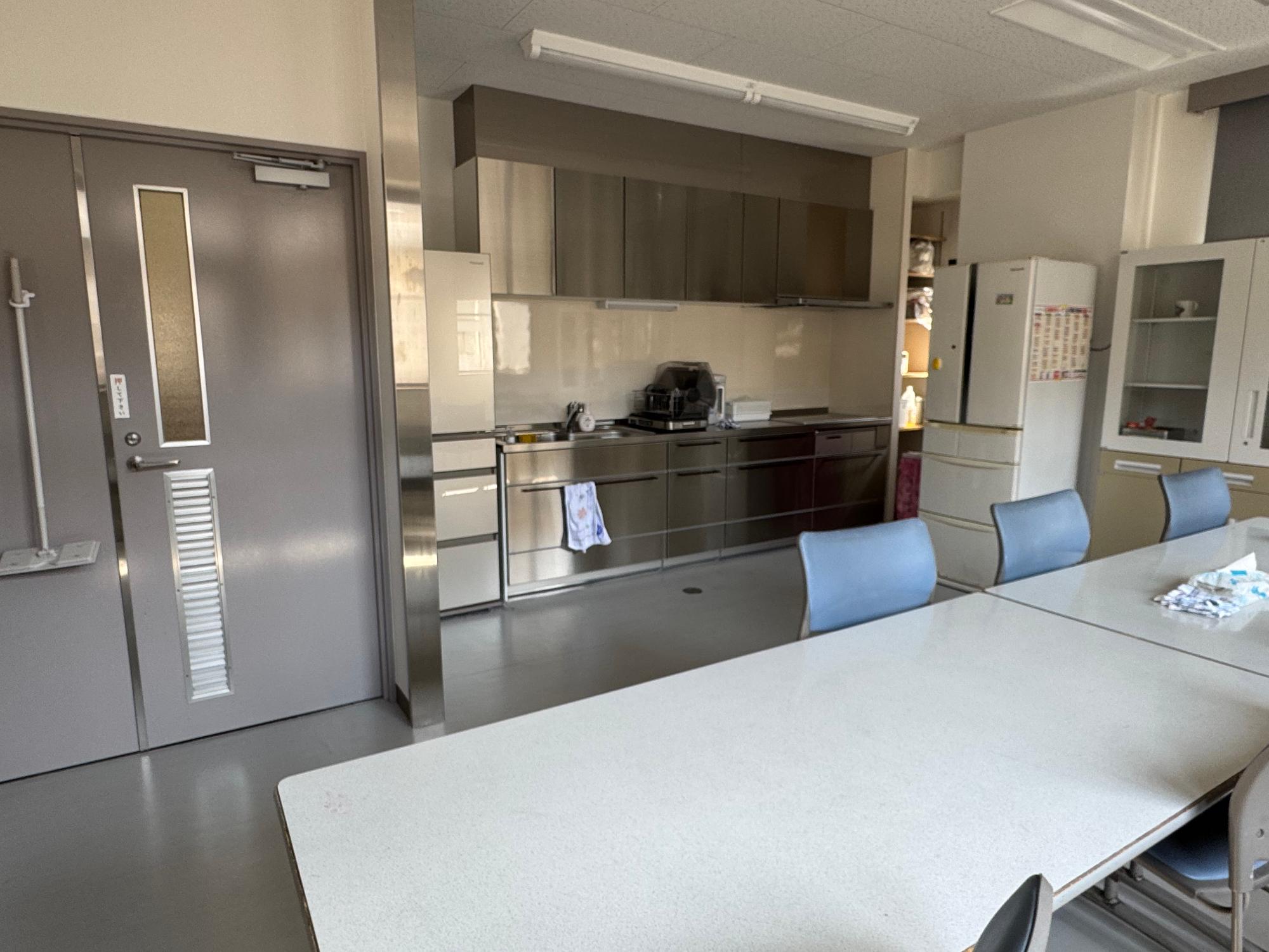 食堂の写真 部屋の真ん中に机が並べられ壁に沿って調理場と冷蔵庫が設置されている