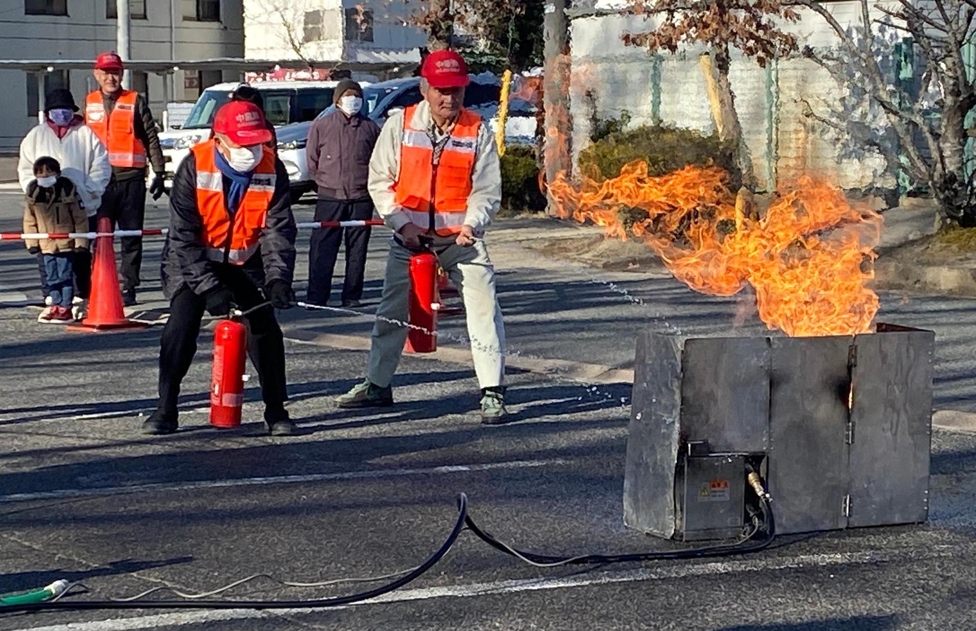 男性二人が火が出る装置に向かって水消火器による消火を行っている様子